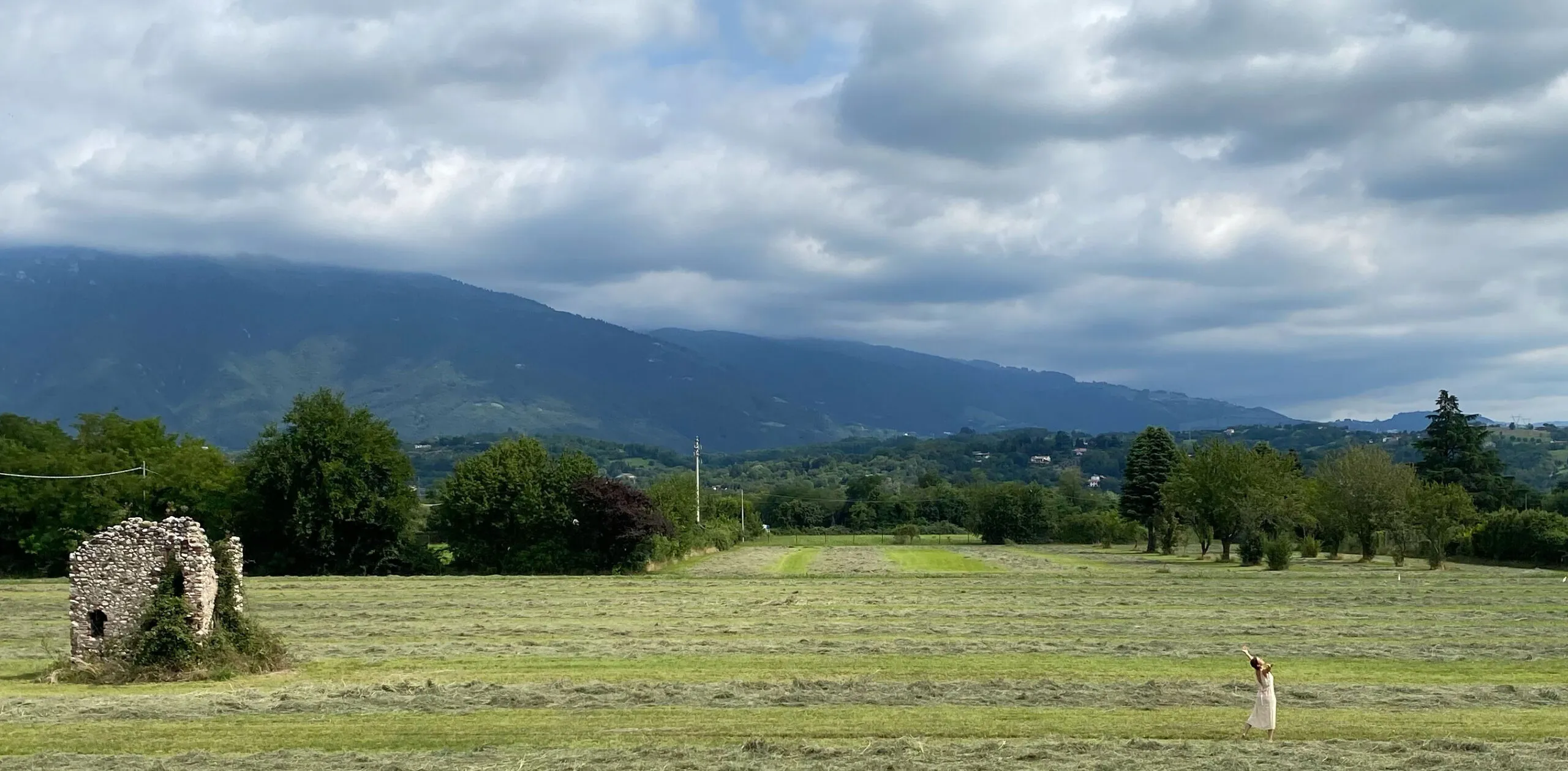 Barbara, piccola in fondo a un prato appena falciato, braccia alzate sotto un cielo di nuvole e montagne